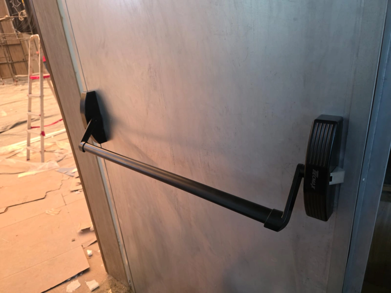 Detalhe de barra antipânico preta em porta de aspecto metálico.