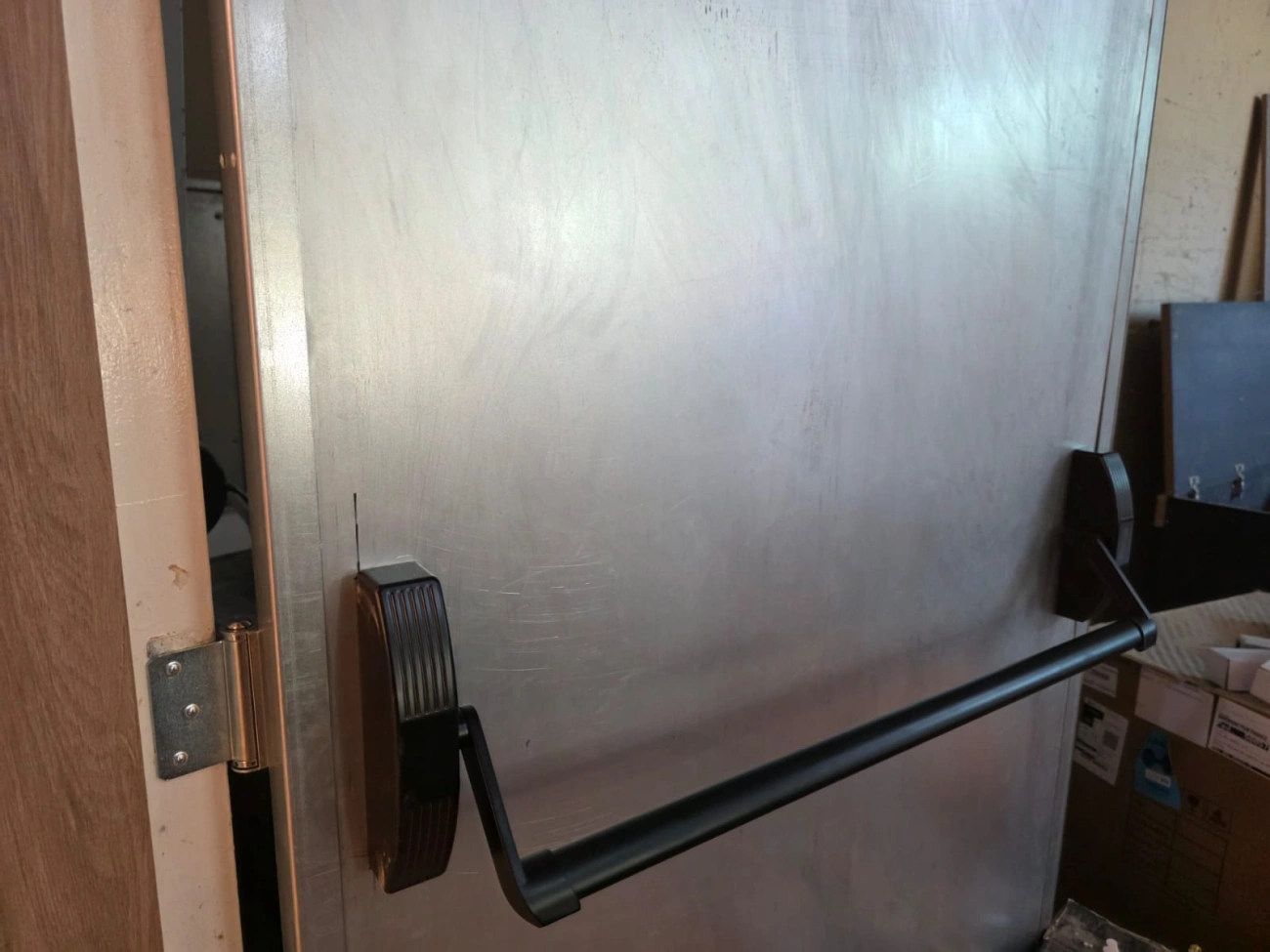 Barra antipânico preta e dobradiça em porta de aspecto metálico.